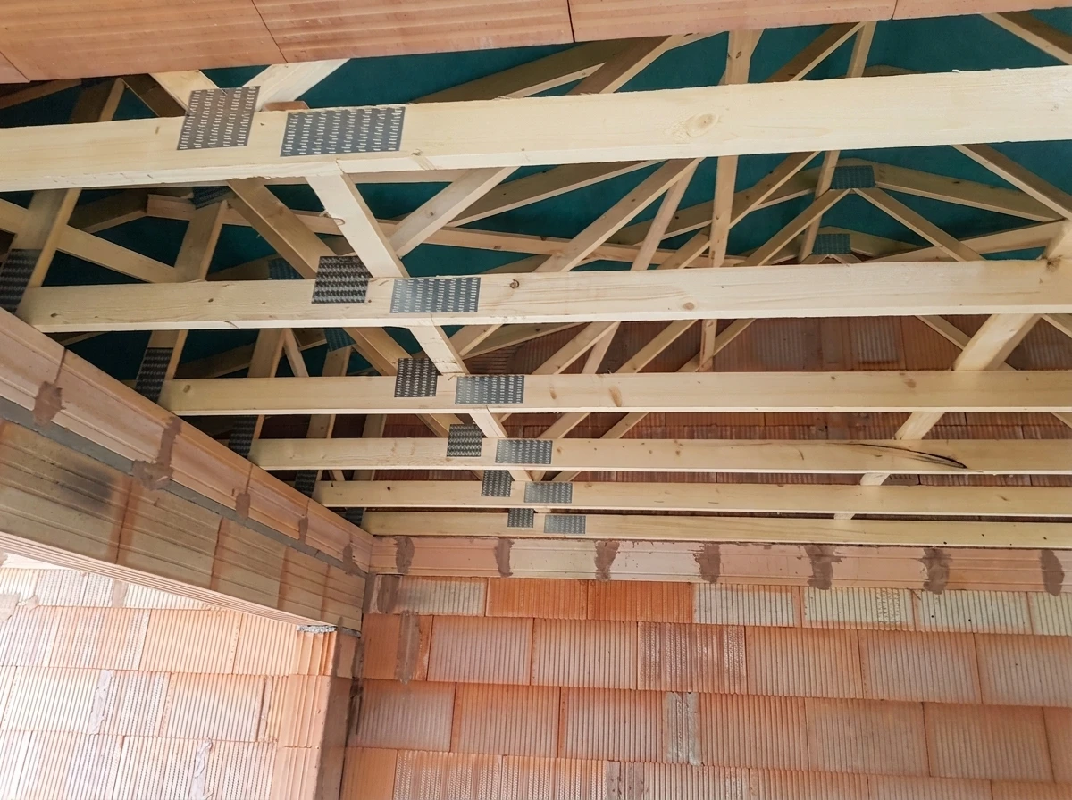 Gros plan sur une structure de charpente en bois massif dur, montrant des poutres robustes assemblées, avec détails des encastrements et jointures traditionnelles. La lumière naturelle souligne la texture du bois massif et la solidité des assemblages. L'image doit transmettre la force, la stabilité et la qualité artisanale de la charpente structurelle.